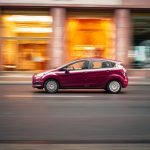Ford Focus czy Fiesta na miasto: co wybrać? Czerwony miejski Ford jedzie szybko po ulicy na tle rozmytego miasta